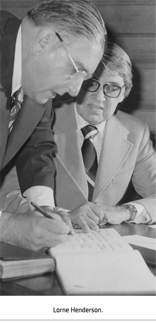 Henderson leaning over a desk and working with the man seated there. Image Caption: Lorne Henderson