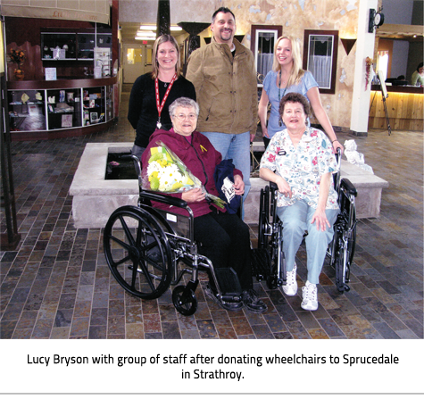 (Lucy and a healthcare worker sit in two wheelchairs in front of three other Sprucedale employees. Image Caption:"Lucy Bryson with group of staff after donating wheelchairs to Sprucedale in Strathroy."), link.