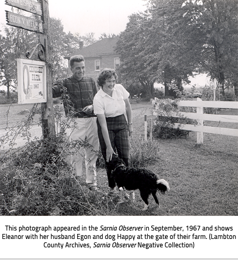 (Eleanor and Egon stand at the front of their property beside a sign that reads "Century Farm 1967". Eleanor reaches down to pet a dog. Image Caption:"This photograph appeared in the Sarnia Observer in September, 1967 and shows Eleanor with her husband Egon and dog Happy at the gate of their farm. (Lambton County Archives, Sarnia Observer Negative Collection)"), link.