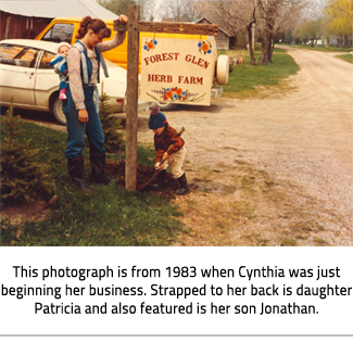 (Cynthia, with her daughter on her back, puts up the "Forest Glen Herb Farm" sign with her young son.Image Caption: "This photograph is from 1983 when Cynthia was just beginning her business. Strapped to her back is daughter Patricia and also featured is her son Jonathan."), link.