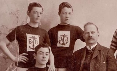A sepia-toned photograph on a brown piece of paper labelled Boys Brigade with Mr. Nisbet. The photograph features eight boys in gym uniforms posing around Nisbet who wears a suit and sits with a basketball.