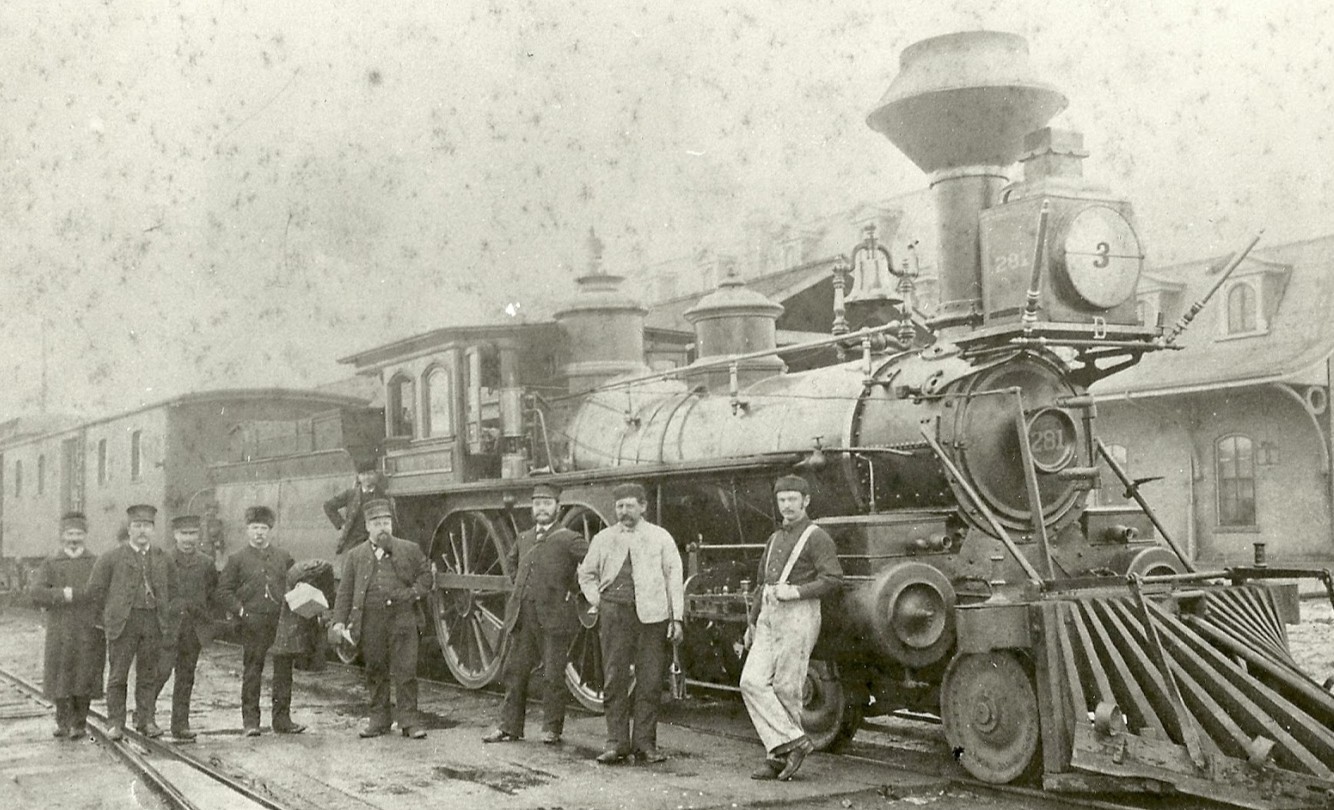 A vintage photograph shows a group of men standing beside a steam locomotive numbered 281 at a train station, with train cars extending into the distance.