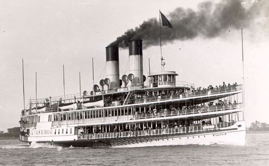 A vintage photograph of the steamboat Tashmoo sailing on the water, filled with passengers on multiple decks and emitting smoke from its twin smokestacks.