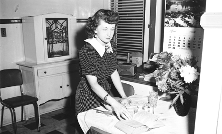 Black and white image of a lady standing in a kitchen at a table.