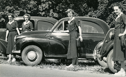 winnifred james among three other women, standing of to the side of the road in front of old cars.