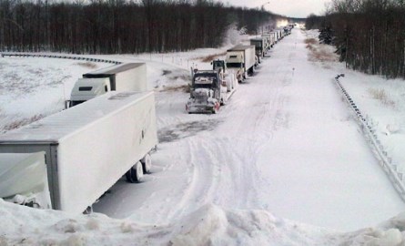 Transport trucks and cars lined up on the side of a road in a snowstorm.