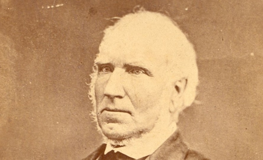 A faded studio portrait of an elderly man in formal attire, seated with one arm resting on an ornate wooden table, gazing slightly off-camera.