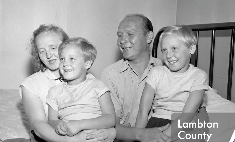 A black-and-white photograph of a smiling family of four, including two young children, sits closely together on a bed in a modest room.