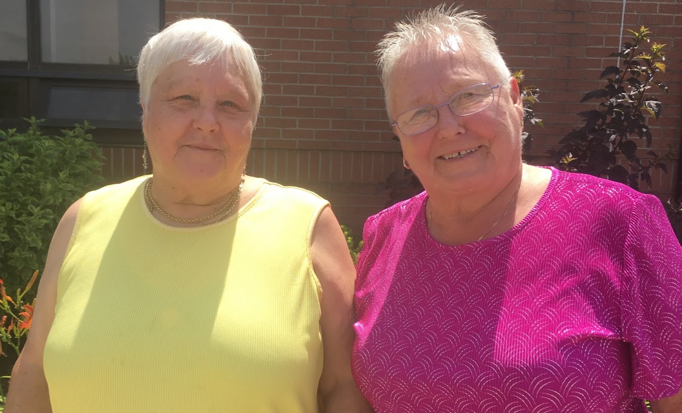 Two smiling older women stand outdoors in front of a brick building and garden, one wearing a yellow sleeveless top and the other wearing a bright pink patterned shirt.