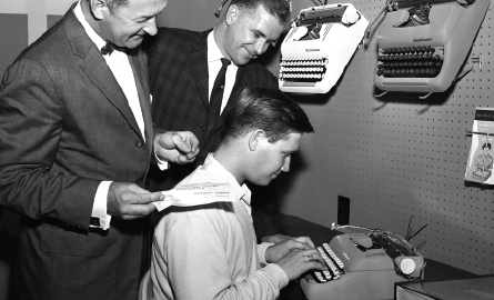 Two men standing over man typing on a typewriter.