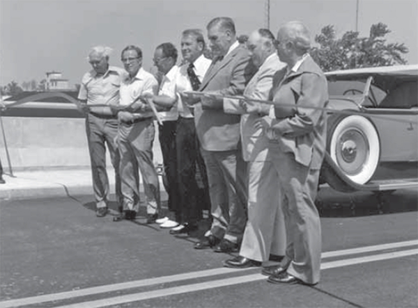 7 men stand behind a ribbon for the Overpass Ribbon Cutting.