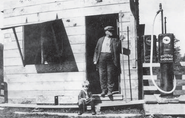 A man leans on the doorway and a child sits on a step of Fenner’s gas station.