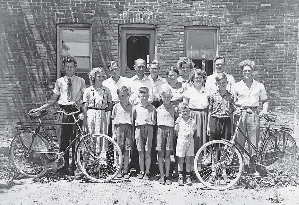 Boere family of 15 stand with bikes in front of brick building.