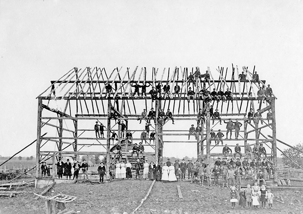 Many people on and around the frame of a large barn.