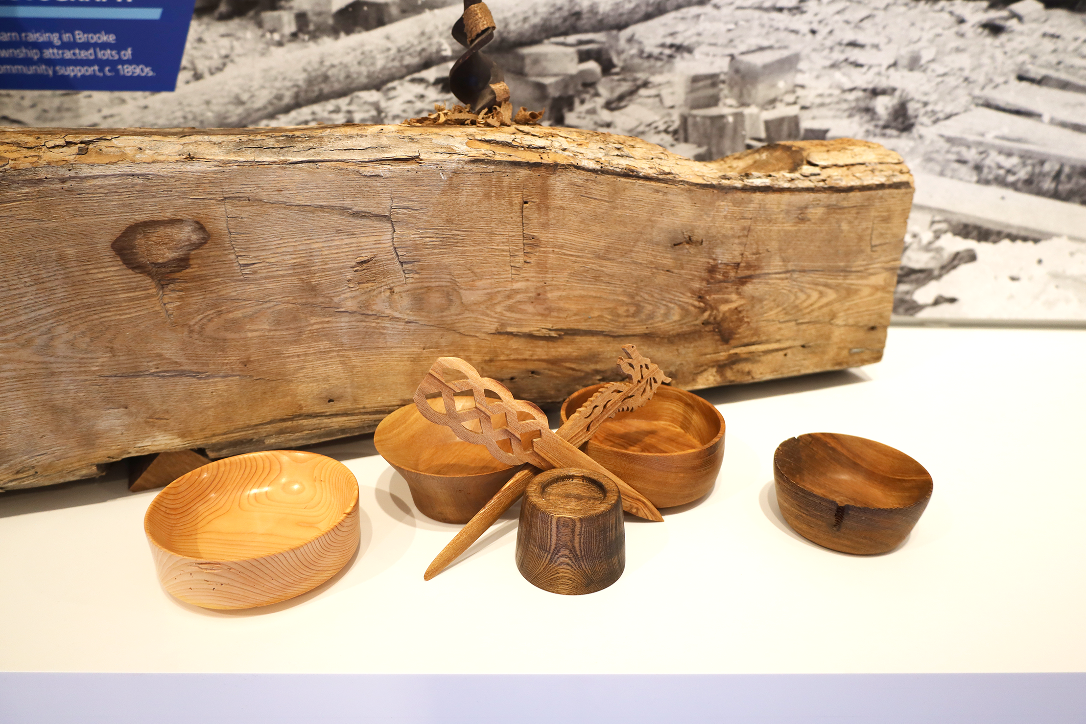 Wooden bowls, candlestick holders, and letter openers of different shapes, sizes, and woodgrain colours, sit in front of a raw wood log.