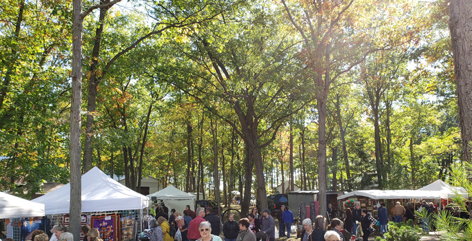 visitors and vendor tent canopies are seen beneath autumn oak trees.