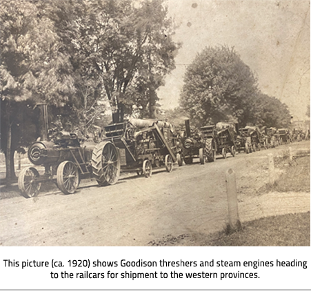 Sepia toned photo of threshers lined up along a country road. Image Caption:This picture (ca. 1920) shows Goodison threshers and steam engines heading to the railcars for shipment to the western provinces. 