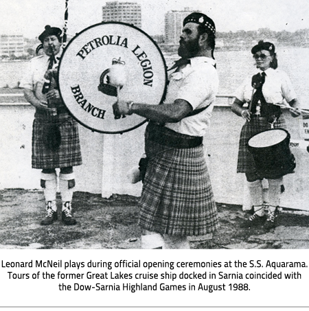 Leonard McNeil playing a large drum that says "Petrolia Legion Branch". He wears a kilt, knee socks, and Scottish hat. In the background, one person plays a bagpipe and another also plays a drum. They are all dressed the same. Image Caption: Leonard McNeil plays during official opening ceremonies at the S.S. Aquarama. Tours of the former Great Lakes cruise ship docked in Sarnia coincided with the Dow-Sarnia Highland Games in August 1988.