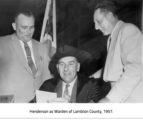 Henderson wears a skewed, wide brimmed hat, and sits at a desk reading a paper; two men read over his shoulder. Image Caption: 