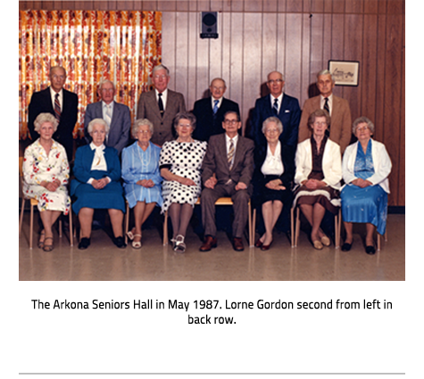(A group of older people posing for a picture. All but one of the men stand in the back while the women sit in front. Everyone is dressed formally.Image Caption: "The Arkona Seniors Hall in May 1987. Lorne Gordon second from left in back row."), link.