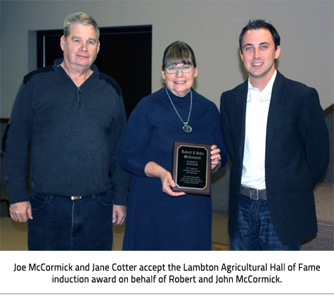 3 people posing for a picture, woman in the middle holds a plaque. Image Caption:Joe McCormick and Jane Cotter accept the Lambton Agricultural Hall of Fame induction award on behalf of Robert and John McCormick.