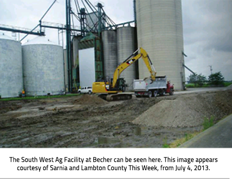  A yellow excavator and a truck in front of a group of silos and grain bins. Image Caption: The South West Ag Facility at Becher can be seen here. This image appears courtesy of Sarnia and Lambton County This Week, from July 4, 2013. 