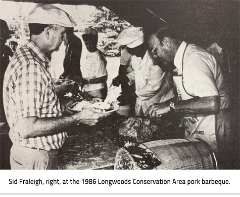 (Men serving meat on barbeque to a line of people. Image Caption: "Sid Fraleigh, right, at the 1986 Longwoods Conservation Area pork barbeque."), link.
