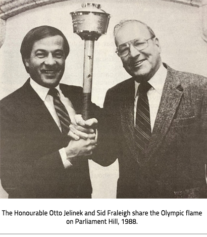 (Two men hold up the Olympic torch between them. Image Caption: "The Honourable Otto Jelinek and Sid Fraleigh share the Olympic flame on Parliament Hill, 1988."), link.