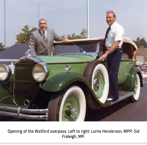 (Two men standing on the standing on the steps on an old green car. Image Caption: "Opening of the Watford overpass. Left to right: Lorne Henderson, MPP; Sid Fraleigh, MP."), link.