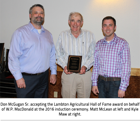Three men pose for photo, the man in the middle holds a plaque. Image Caption:Don McGugan Sr. accepting the Lambton Agricultural Hall of Fame award on behalf of W.P. MacDonald at the 2016 induction ceremony. Matt McLean at left and Kyle Maw at right.