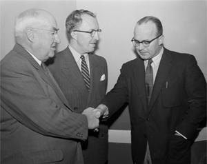 Three men all in suits and wearing glasses talking, two of the men shake hands.