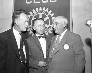 Three men all in suits in front of a "Rotary International" banner. The three men wear pins that say "Rotary International" on them.