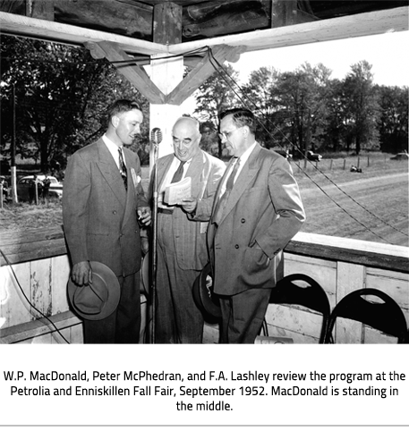 Three men in suits stand around a microphone, the one in the middle reads off a book. Image Caption: : W.P. MacDonald, Peter McPhedran, and F.A. Lashley review the program at the Petrolia and Enniskillen Fall Fair, September 1952. MacDonald is standing in the middle. 
