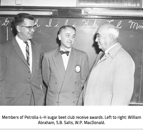 Three men stand together talking in front of a chalkboard.  Image Caption: Members of Petrolia 4-H sugar beet club receive awards. Left to right: William Abraham, S.B. Salts, W.P. MacDonald.