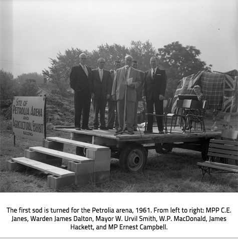 MacDonald stands on a wagon being used as a stage and read a booklet in front of a microphone. Behind him stands a group of men. To the left a sign reads "Site of Petrolia Arena and Agricultural Exhibit Building. Image Caption:The first sod is turned for the Petrolia arena, 1961. From left to right: MPP C.E. Janes, Warden James Dalton, Mayor W. Urvil Smith, W.P. MacDonald, James Hackett, and MP Ernest Campbell.
