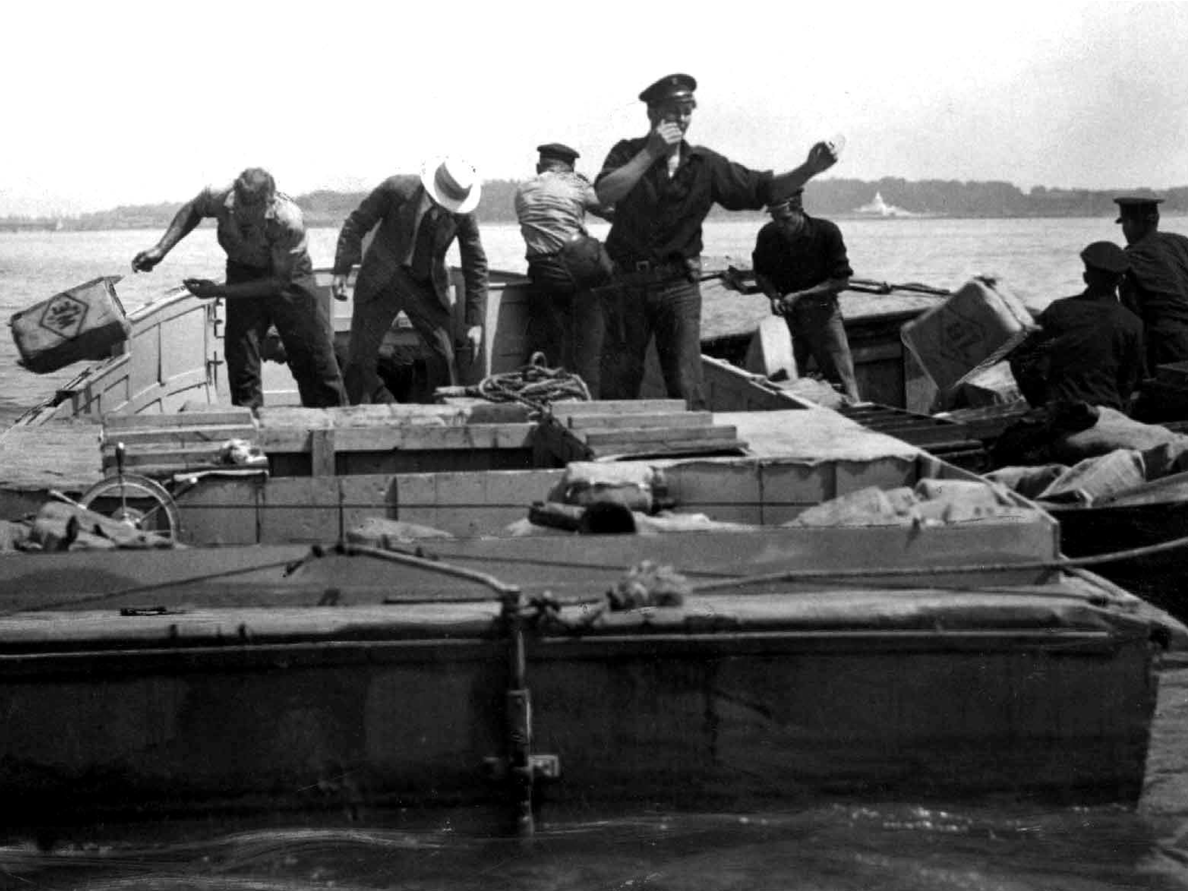 Black-and-white photo of several people loading crates onto boats on open water.