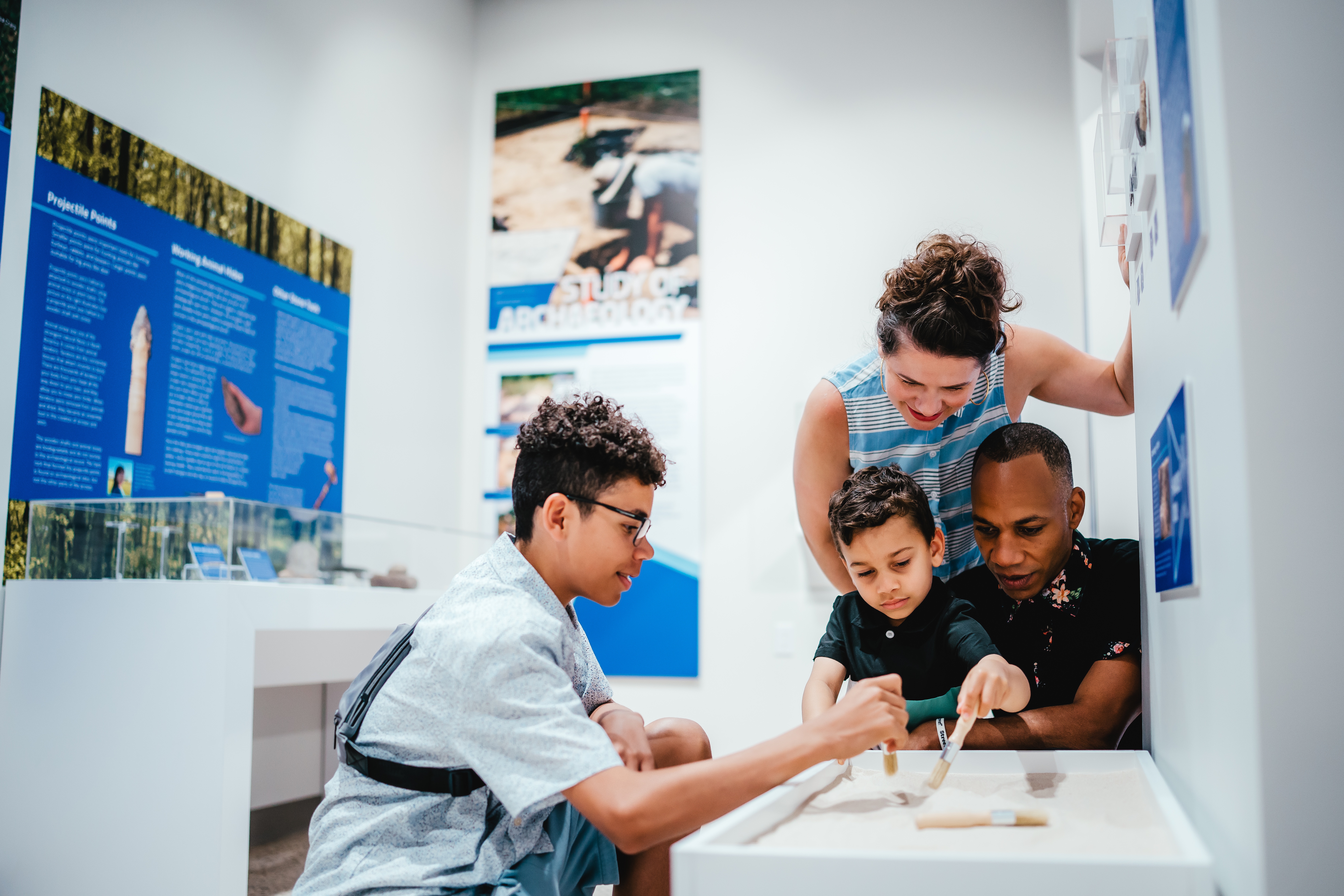 A family of four using brushes to find artifacts in a sand table