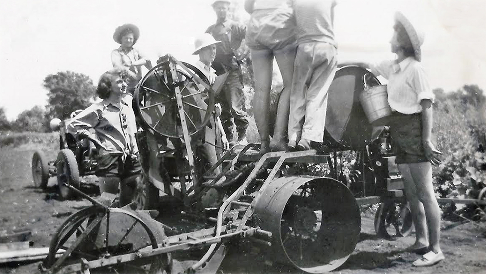Farmerettes and farmers on farm machinery.