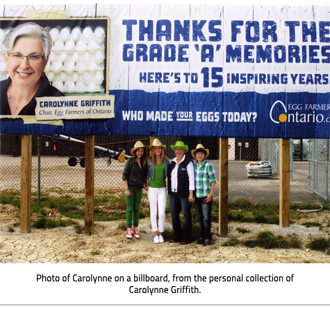 (Road sign with Carolynne Griffith on it advertising their Egg Farm. Family members stand together underneath it.The sign reads "Carolynne Griffith", "Chair, Egg Farmers of Ontario", "Thanks for the grade 'A' memories. Here's to 15 inspiring years", "Who made your eggs today? Egg Farmer Ontario.ca". Image Caption: "Photo of Carolynne on a billboard, from the personal collection of Carolynne Griffith"), link.