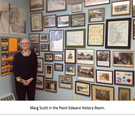 (Older woman standing beside two walls covered with framed pictures, maps, and documents. Image Caption: "Marg Scott in the Point Edward History Room."), link.