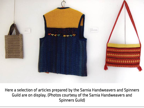 (A woven beige bag, an orange and blue vest, and a patterned red and yellow purse hanging on a white wall. Image Caption: "Here a selection of articles prepared by the Sarnia Handweavers and Spinners Guild are on display. (Photos courtesy of the Sarnia Handweavers and Spinners Guild)"), link.