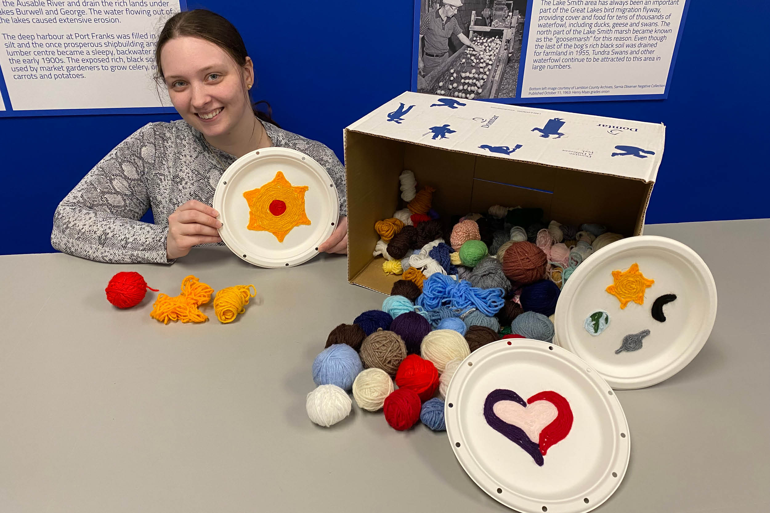 Lambton Heritage Museum staff member Karissa Ramsey poses with a box of yarn balls and a yarn painting of a sun.