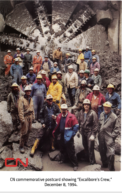 (A crew of men wearing hard hats posing for a photo in front of a tunnel. In the bottom left corner is the red 'CN' logo. Image Caption: "CN commemorative postcard showing “Excalibore’s Crew,” December 8, 1994."), link.