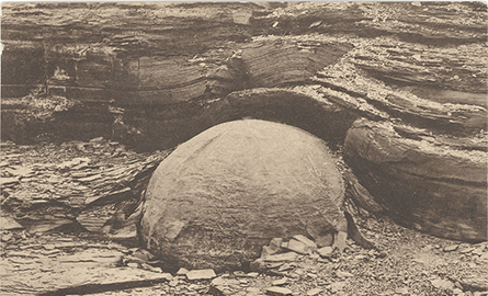 A sepia toned post card showing a large sphere (kettle) breaking free of the ground and stone surrounding it. The text at the bottom reads: Kettle and Shale at Kettle Point, Near Forest, Ontario, Canada. Image Caption: This postcard shows a kettle in situ. You can see how the layers of the surrounding shale bent around the spherical kettle as it grew. 