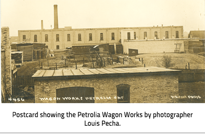 (Wood piles by brick buildings with text. Image Caption: "Postcard showing the Petrolia Wagon Works by photographer Louis Pesha."), link.