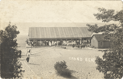 (A postcard of a large crowded pavilion on Grand Bend beach. To the left of it there is a shed, and below there is the text "GRAND BEND".), link.