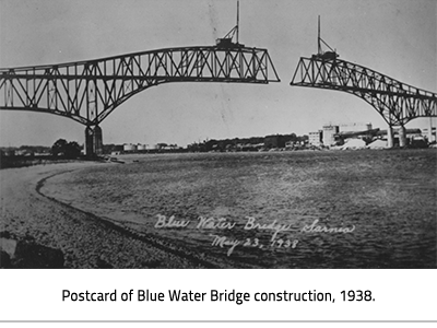 (Black and white postcard of the two halves of the Blue Water bridge being built over the St.Clair River. A crane sits a top each half. The cursive text on the bottom of the card reads, "Blue Water Bridge Sarnia May, 23 1938". Image Caption: "Postcard of Blue Water Bridge construction, 1938."), link.