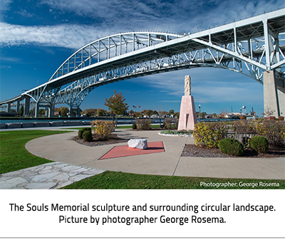 (A tall, reddish stone monument in the middle of a circular garden made up of hedges and bushes. In the background is the Bluewater Bridge over the St. Clair River. Image Caption: "The Souls Memorial sculpture and surrounding circular landscape. Picture by photographer George Rosema."), link.