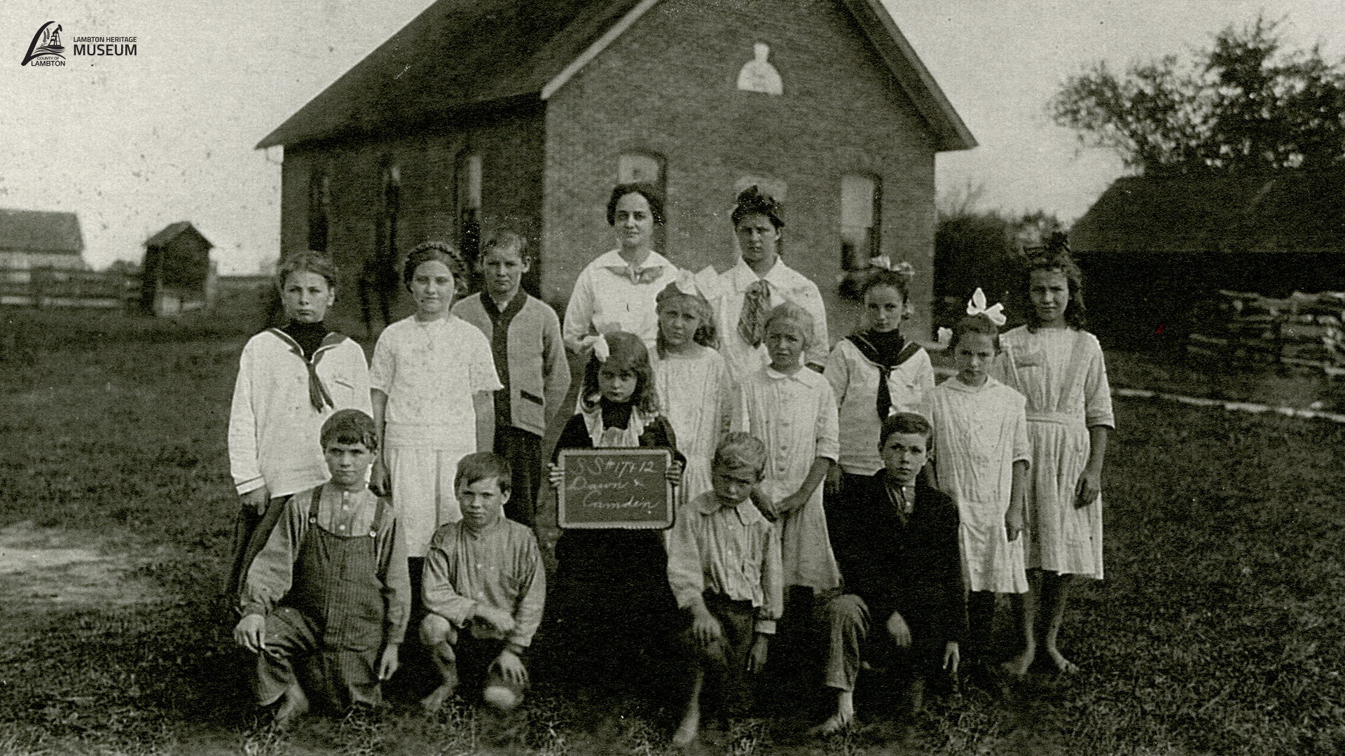 Historic black-and-white photograph of a rural school group, showing a teacher and schoolchildren posed outside a one-room schoolhouse