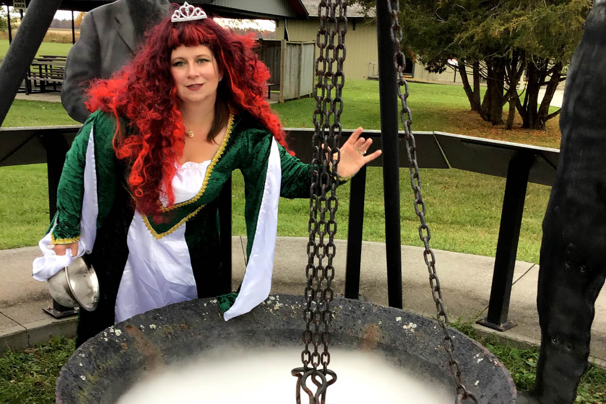Christina Sydorko in a green dress with red wig stands next to a cauldron in an outdoor display.